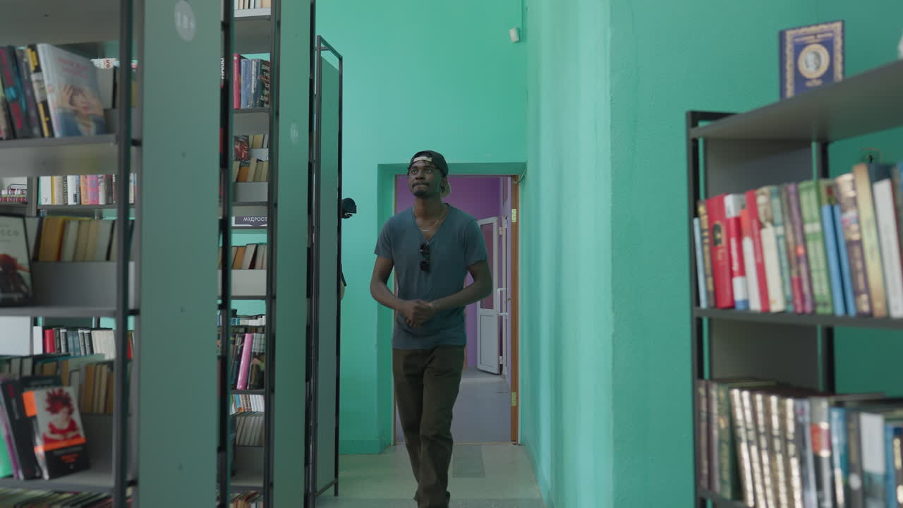 Man enters modern library dancing with playful steps, wearing cap and gray shirt, walking between tall shelves of colorful books, joyful mood, casual style, study space, academic vibe