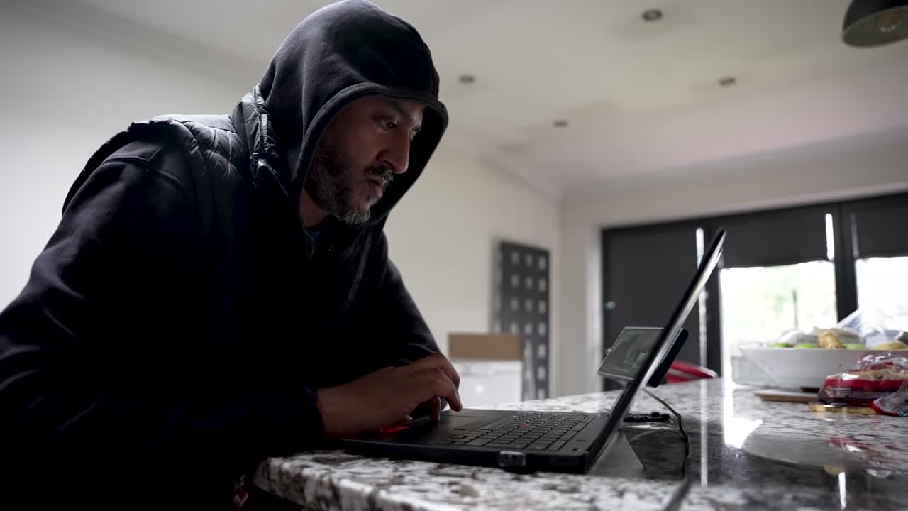 A South Asian man wearing a hoodie is working on a laptop at a kitchen table. A home office setup for remote work and online communication