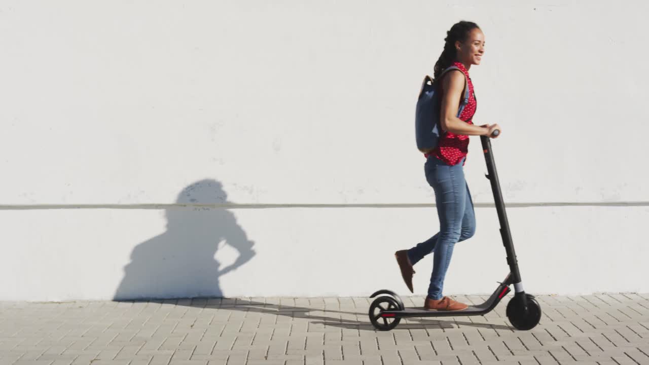 African american woman wearing backpack riding scooter on promenade by the sea