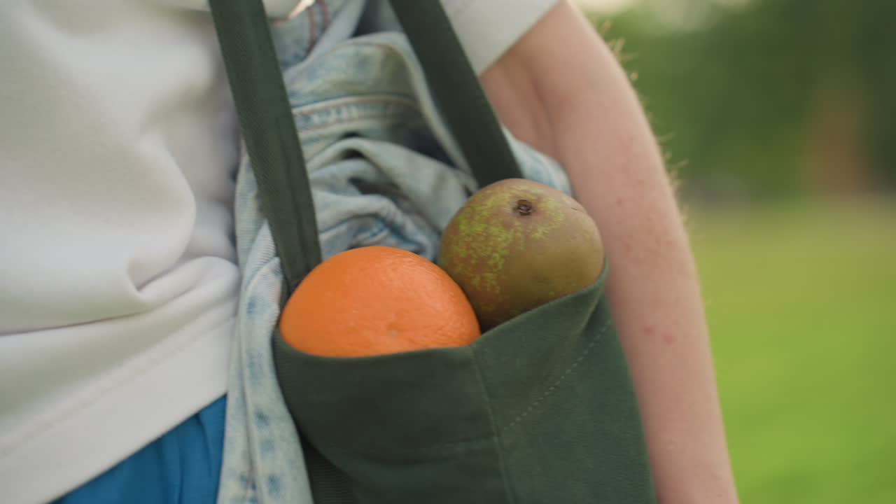 Hand view of caregiver carrying fruits in black canvas bag while walking, denim fabric and white shirt visible, bright daylight highlighting orange and pear, healthy outdoor movement