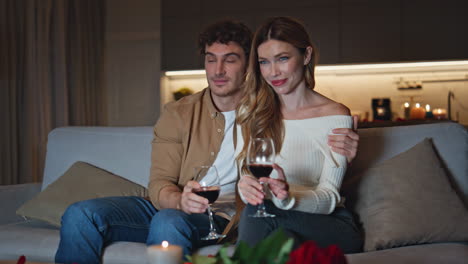 gentle lovers sitting couch watching tv with wine glasses. man hugging woman