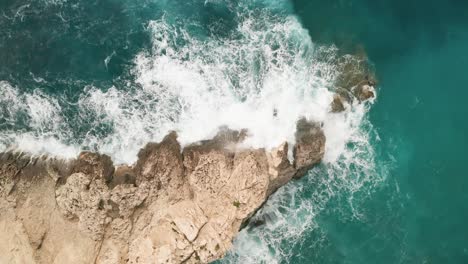 Viewed-from-an-overhead-drone,-the-scene-unveils-striking-turquoise-waters,-with-white-waves-crashing-against-a-vibrant-brown-rock-along-Gozo's-coast,-sending-sea-spray-soaring