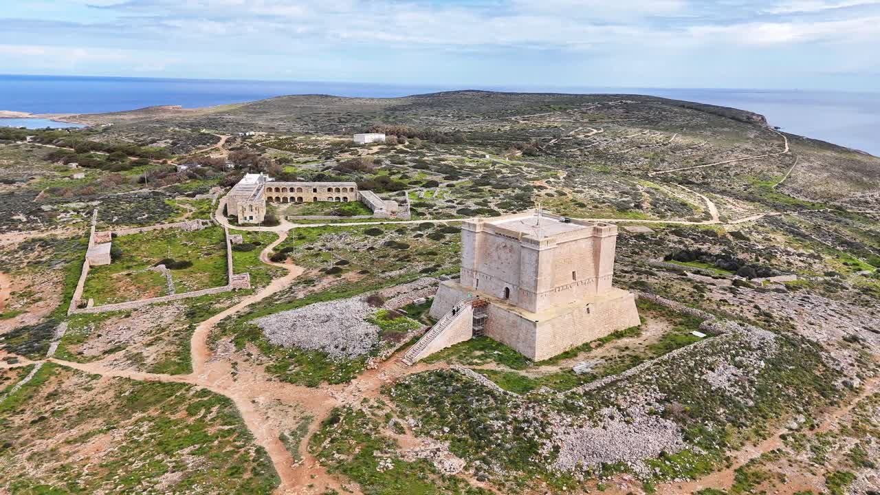 Malta, Comino'daki St Mary Kulesi'nin havadan drone görüntüsü, kayalık bir arazi üzerinde, görünen patikalar ve tarihi yapılarla, gün ışığında sahil şeridine ve açık denize bakıyor.