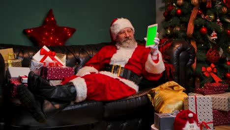 santa claus lies on the sofa near decorated christmas tree and gift boxes, shows tablet with a green screen chrome key, making thumb up gesture. new years advertising, promotion. slow motion