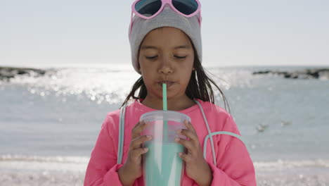 portrait of little girl drinking beverage on beach wearing beanie and pink clothes