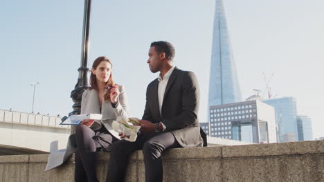 Two-millennial-colleagues-take-a-break-on-the-embankment-eating-and-talking-sitting-on-the-Thames-wall-by-the-river-near-London-Bridge,-low-angle,-close-up