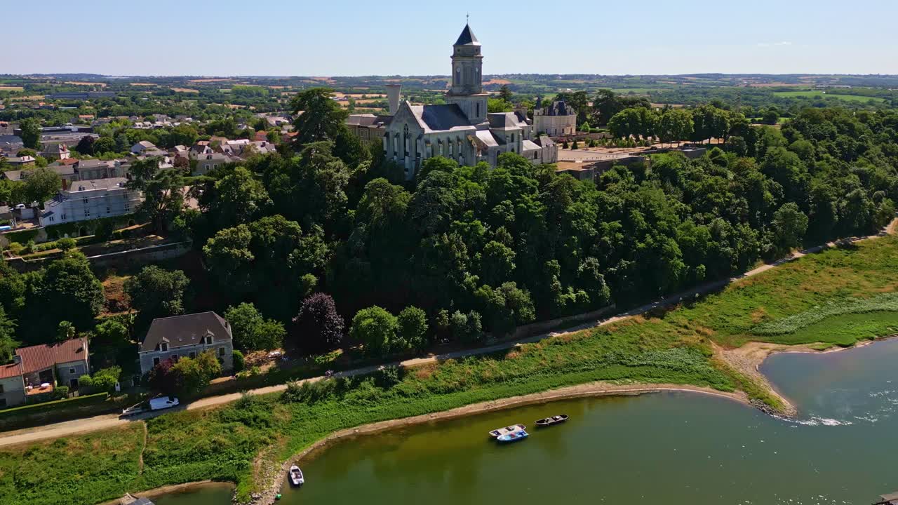 Droneopptak fremover over Loire-elven, passerende båter og trær, som nærmer seg Saint-Florent klosterkirke på en liten høyde med nærliggende hus - Mauges-sur-Loire i Frankrike.
