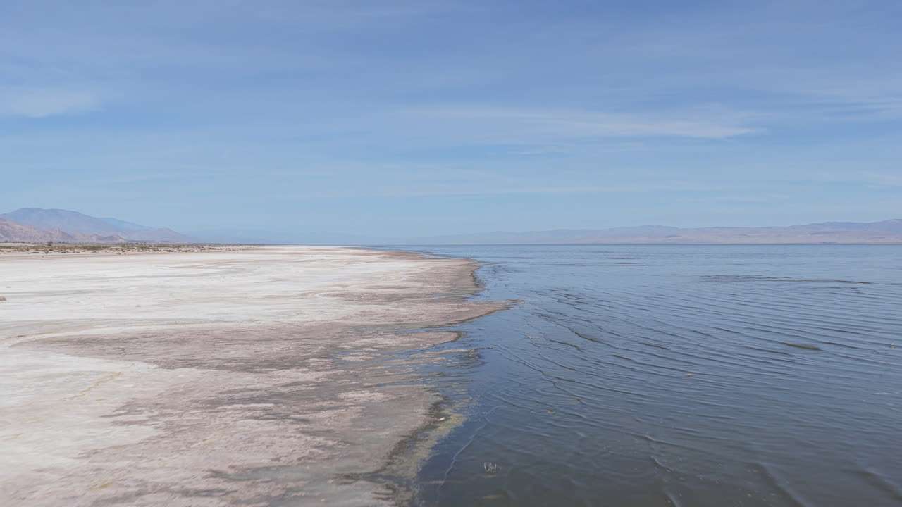 Imagem de drone em baixa altitude voando para a frente ao longo da costa do Mar Salton na Califórnia, com água aberta à direita e a costa em retração e o leito do lago exposto à esquerda.