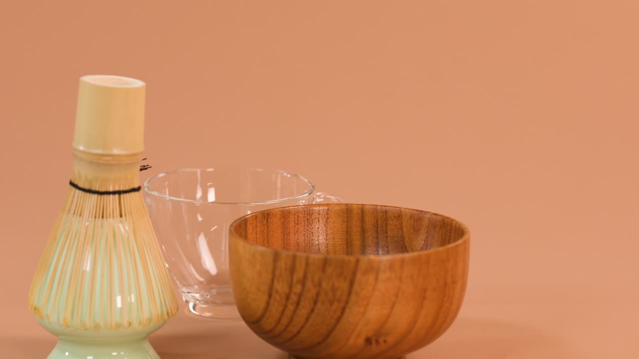Clear water pours from glass pitcher into wooden bowl, bamboo whisk, soft studio lighting