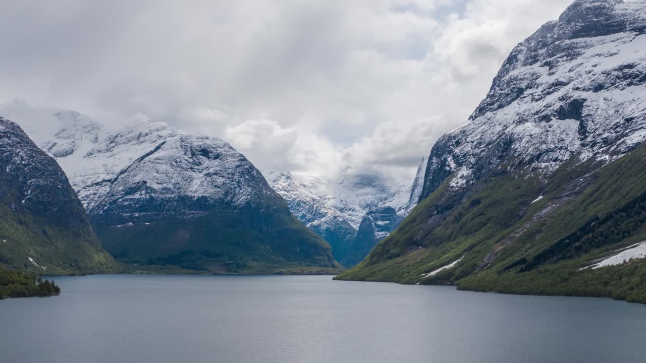 Thick, white clouds whirling above the vast dark waters of the Lovatnet lake, Norway. Ripples running on the water. Mountains tower on the horizon. Forest is covering the shores of the lake.