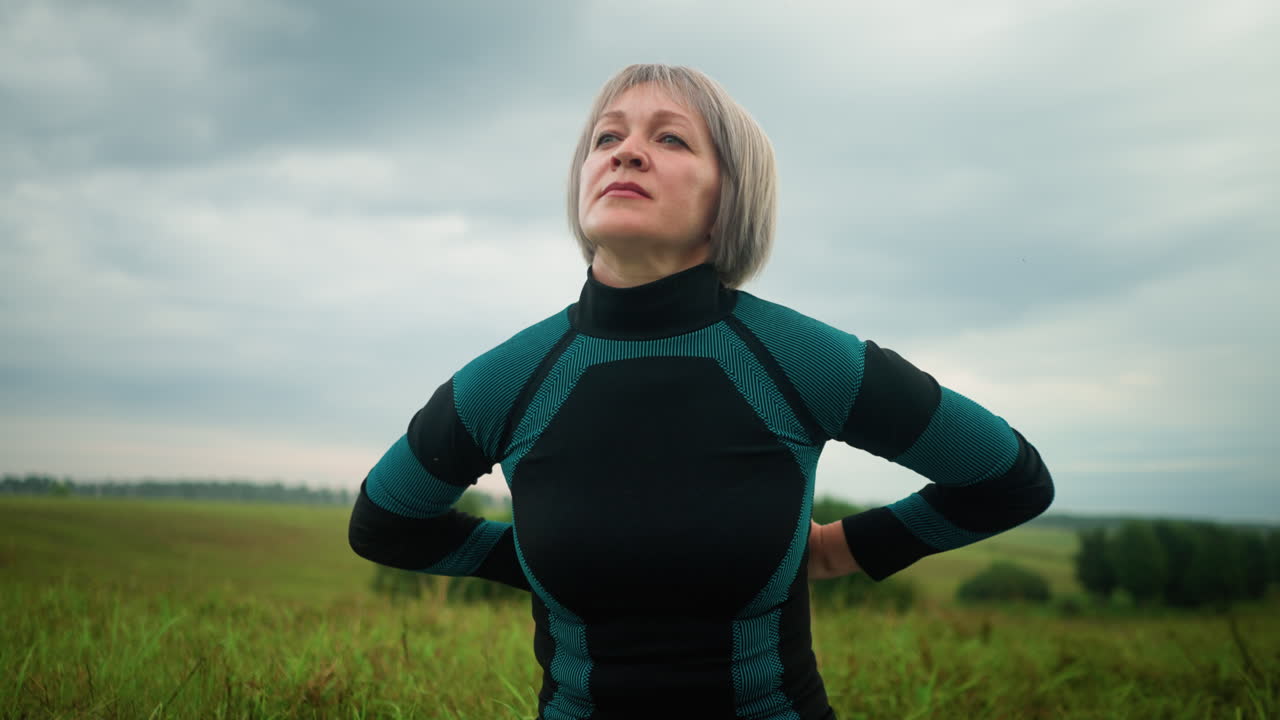 Middle-aged woman sitting in sukhasana pose practicing yoga, gazing into the distance with a calm expression, in the background, blurred trees are lined up under a cloudy sky