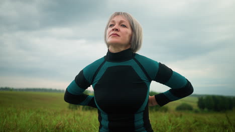 mujer de mediana edad sentada en sukhasana postura practicando yoga, mirando a la distancia con una expresión tranquila, en el fondo, los árboles borrosos están alineados bajo un cielo nublado