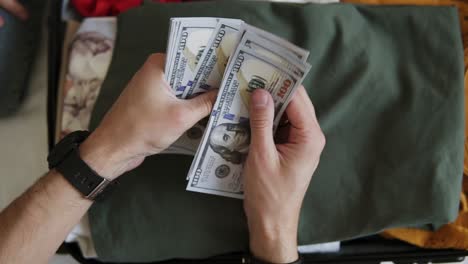 Unrecognizable-man-hands-with-pile-of-money,-US-dollars-notes---counting-under-the-suitcase-with-clothes.-Counting-money-for