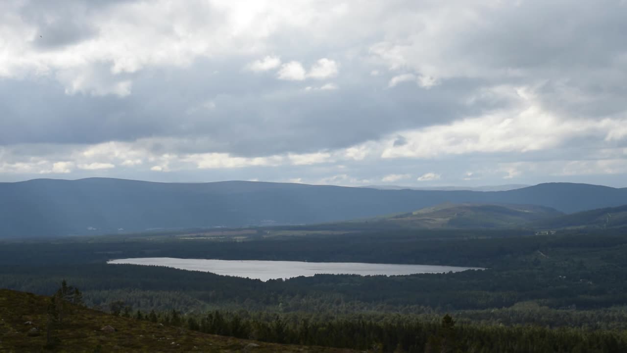 Wide view of peaceful lake nestled in dense forest with distant hills and cloudy sky creating dramatic light in remote northern Scotland, showcasing natural beauty and serene highland landscape