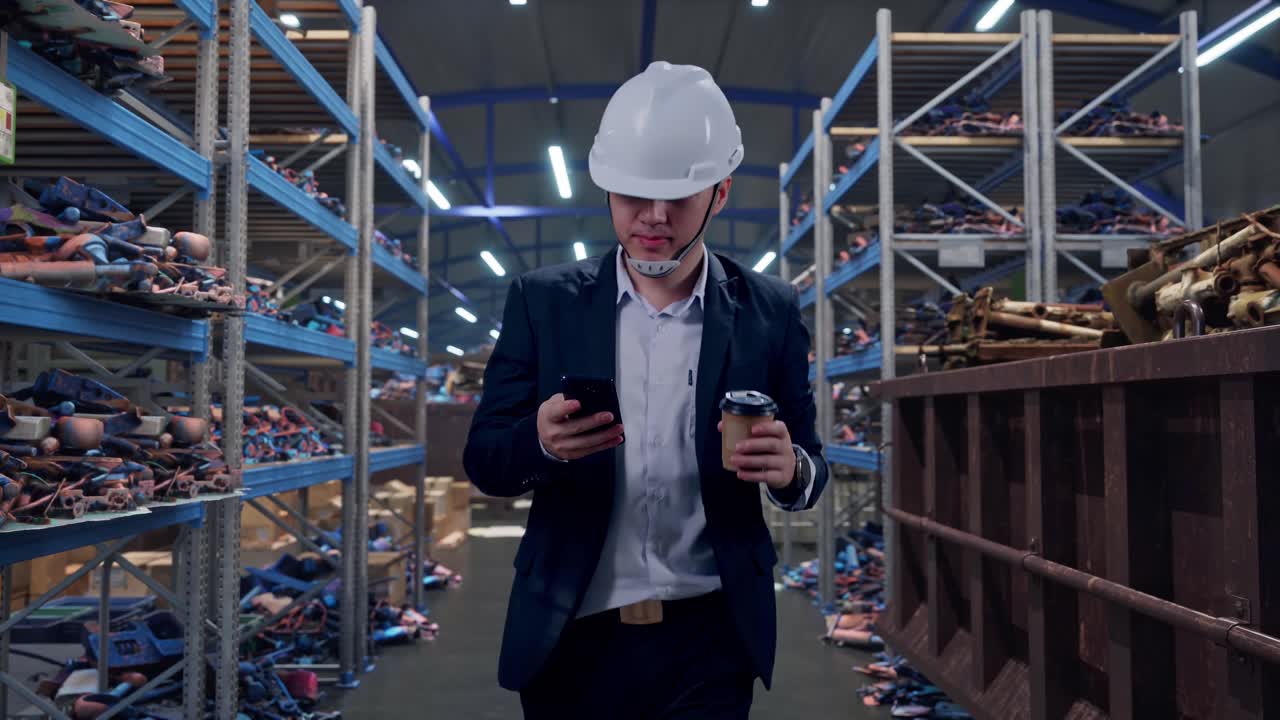 Вид спереди: Азиатский бизнесмен использует смартфон и пьёт кофе, идя по промышленному складу металлолома и переработки.