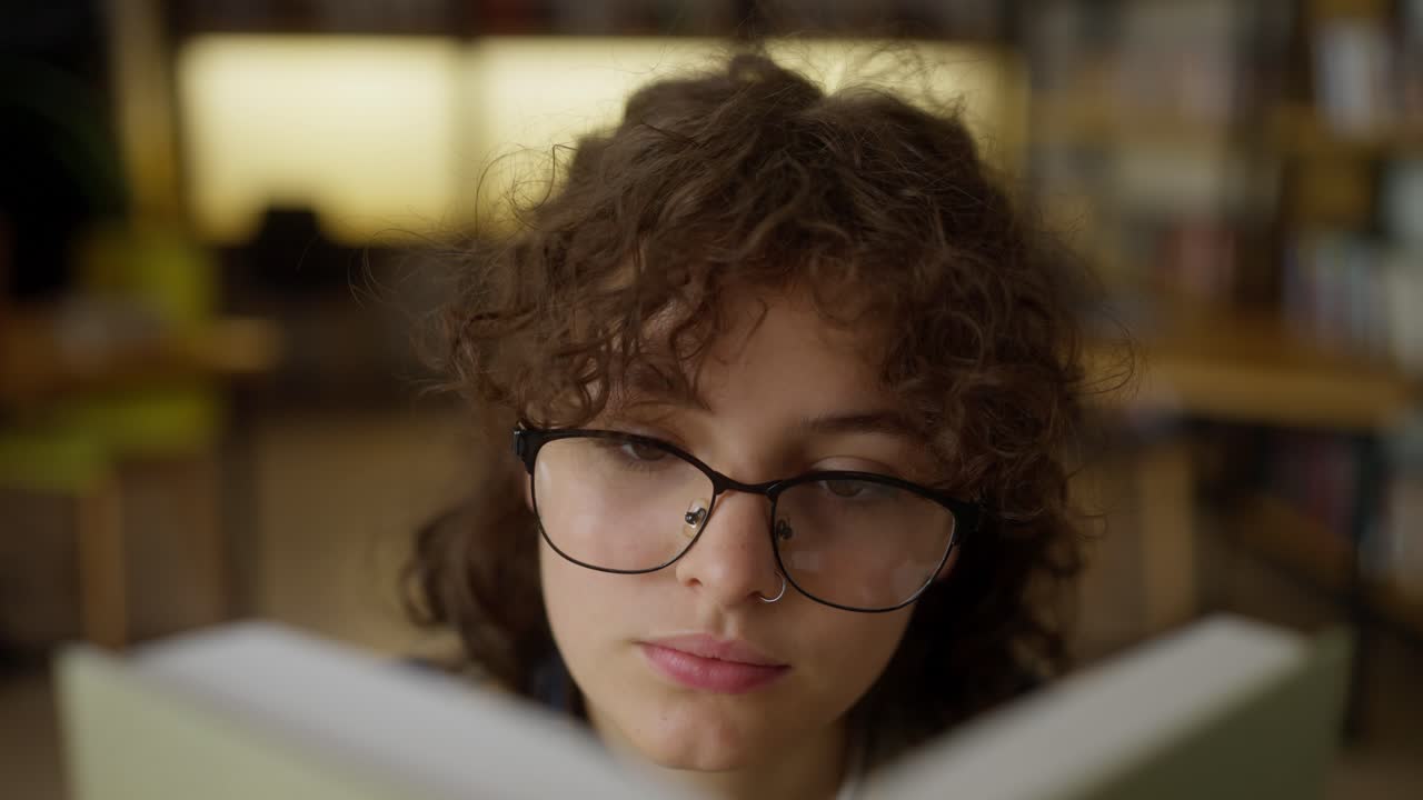 primer plano de una chica con cabello rizado que usa gafas leyendo un libro en una mesa en la biblioteca