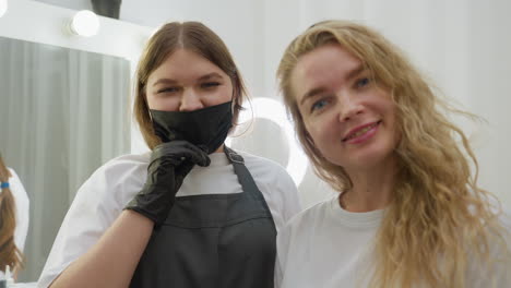 client and beautician stand closely inside salon, both smiling at camera with relaxed expression. beautician wears mask and apron, client has long blonde hair, background shows mirror with lights