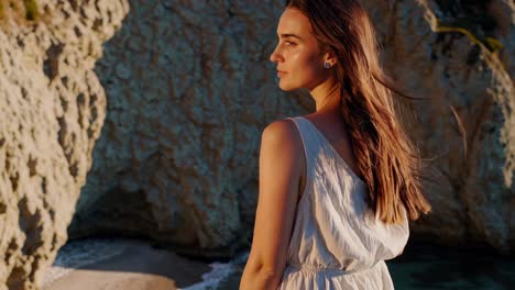 woman in a white dress stands on a clifftop at sunset, her hair blowing in the wind, enjoying the serene view of the ocean and beach below