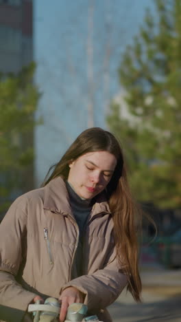una mujer joven con una chaqueta de melocotón y camisa interior gris camina por un parque en un día soleado, con algo en la mano, mientras se sienta en un banco. el fondo incluye vegetación y edificios