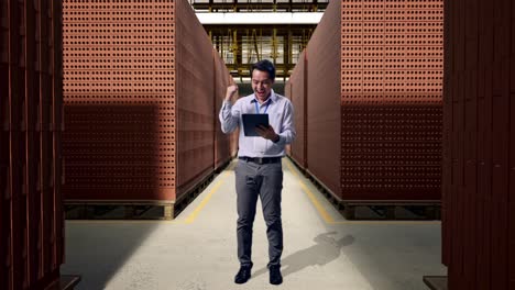 full body of an asian male professional worker standing with his tablet with red brick packed in stacks are stored, he raises his fist up with screaming goal after typed on the tablet