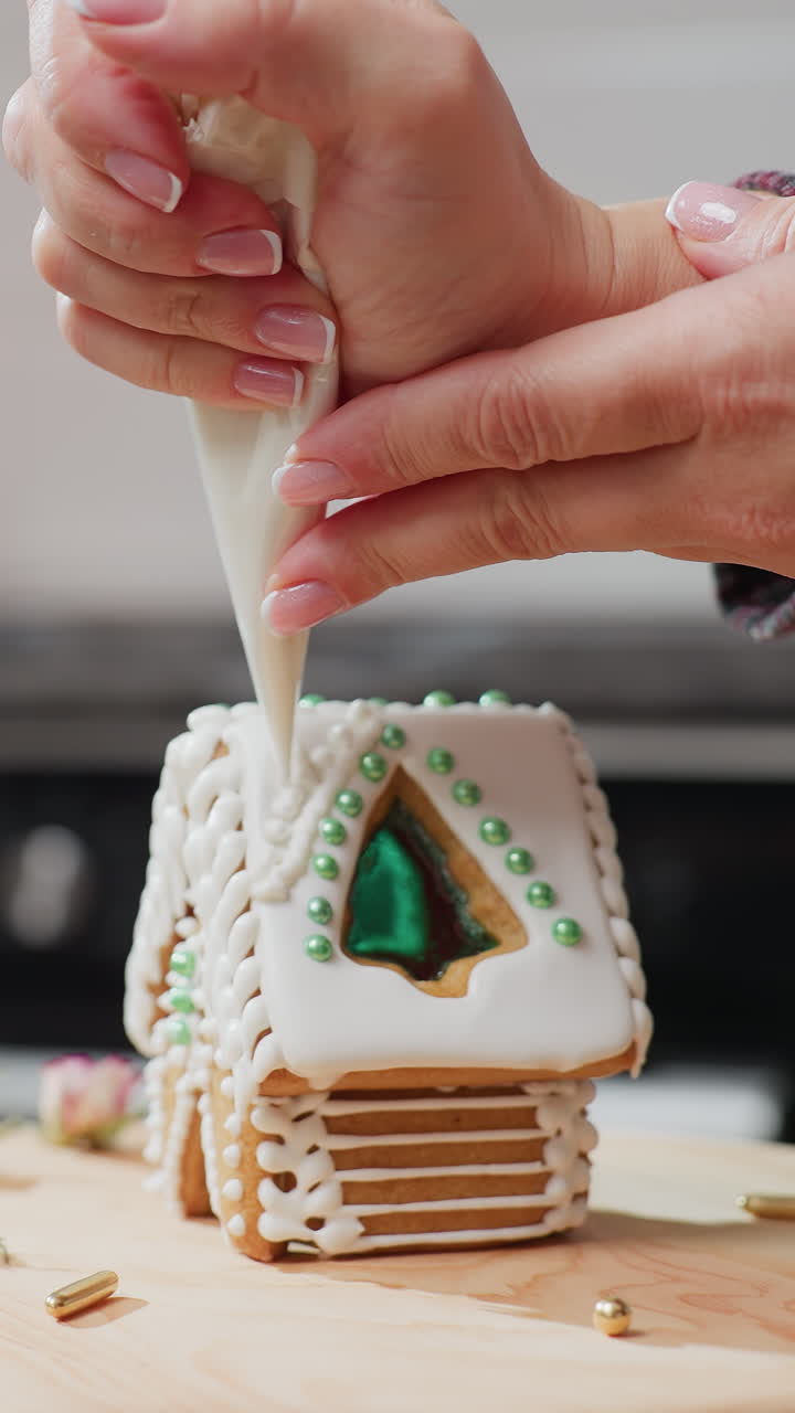 manos aplicando cuidadosamente hielo blanco en la casa de pan de jengibre usando bolsa de tubería, cuentas verdes, flores secas, bandeja de madera y luz de vela cálida