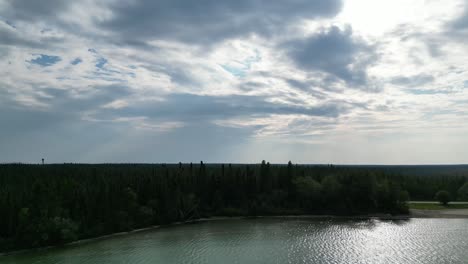 Eine-Bewegte-Luftdrohnenaufnahme-Des-Clearwater-Lake-Vom-Sunset-Beach-Am-Rande-Des-Pas-Gebiets-Im-Sommer-2023-An-Einem-Bewölkten-Tag,-Die-Die-Wolken-Und-Die-Durchscheinende-Sonne-Hervorhebt