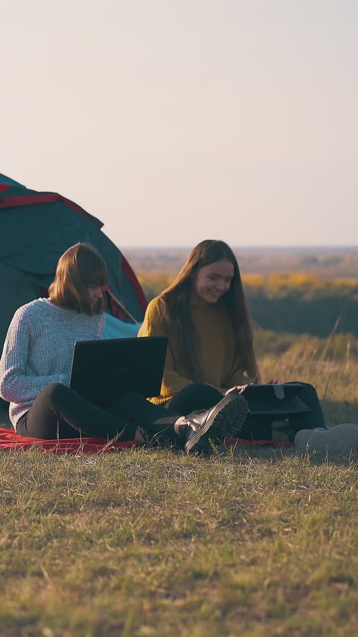 joyful girl hikers with tablet and laptop work on red plaid near tent on steep river bank in warm autumn evening
