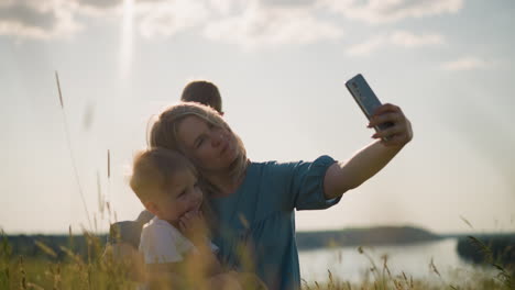 close-up of a woman in a blue dress sitting on grass, holding a smartphone while filming herself and her son sitting on her lap. they both smile as her older son in the background is on his phone