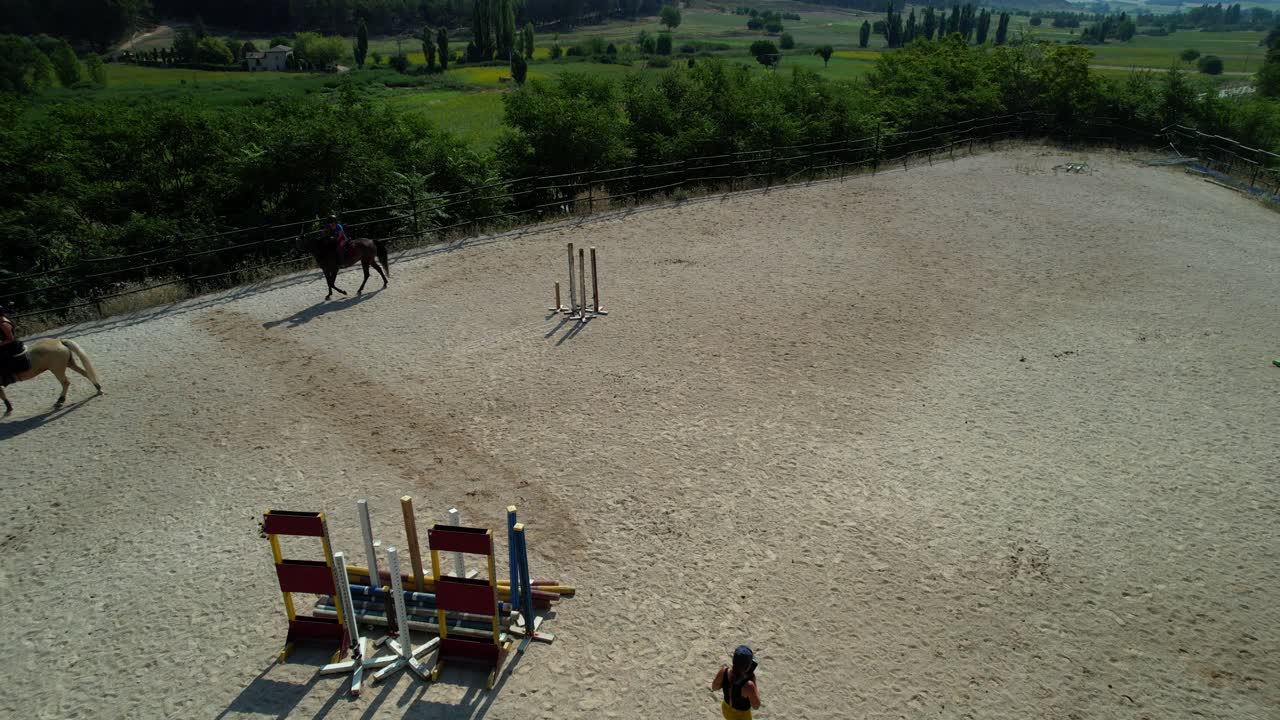 Tilt up drone shot reveals a vast green landscape that surrounds a horse training track with riders and coach at work