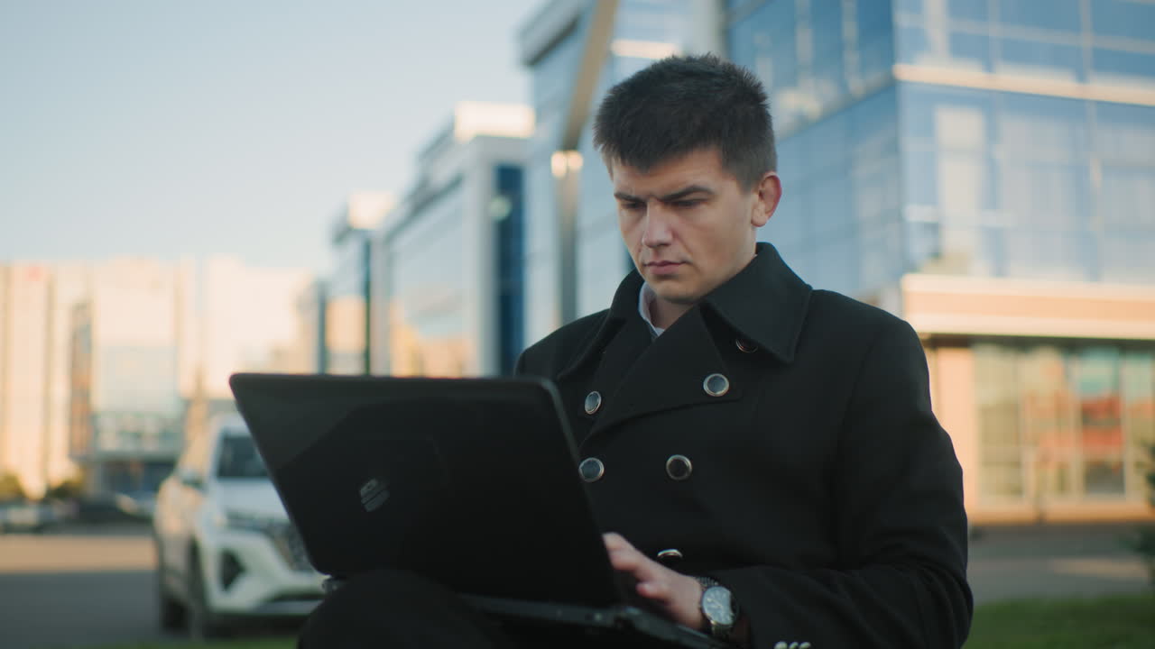 야외에 앉아 다리를 은 채 노트북을 작동하는 은행가, 작업에 집중, 배경에는 주차된 자동차, 현대적인 유리 건물, 그리고 초록색이 있습니다.