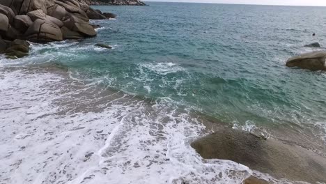 Continuous-wave-rushing-up-to-beach-over-bay-at-corner,-side-overlook-static-dronefly-shot