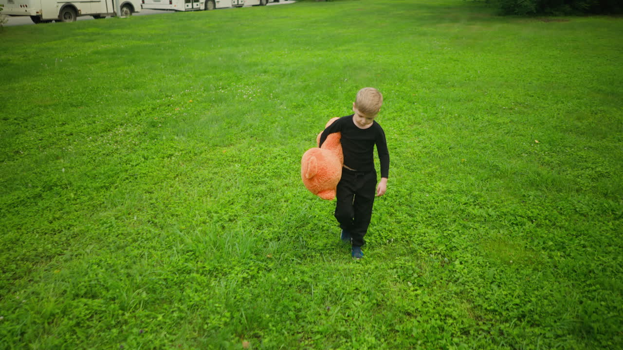 Young boy in black outfit walking across wide grassy field holding large orange teddy bear under arm, approaching from distance with line of white buses and official building visible in background