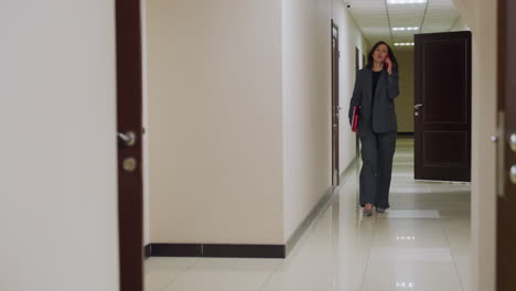 businesswoman walking through office corridor talking on phone, holding documents in formal attire, modern office space, professional work environment, multitasking, and corporate life