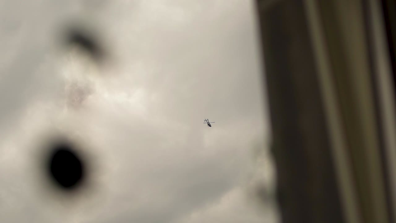 Helicopter flying through a cloudy sky, partially obscured by a window's reflection
