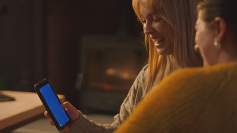 Two-Female-Friends-Relaxing-On-Sofa-Looking-At-Mobile-Phone-With-Cosy-Fire-Holding-Glass-Of-Wine