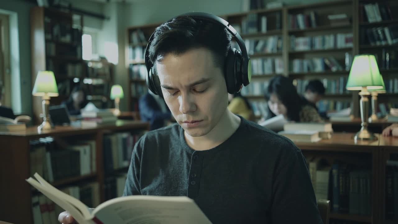 Student iført svart henley og hodetelefoner studerer med en åpen bok på biblioteket, med lamper og hyller.