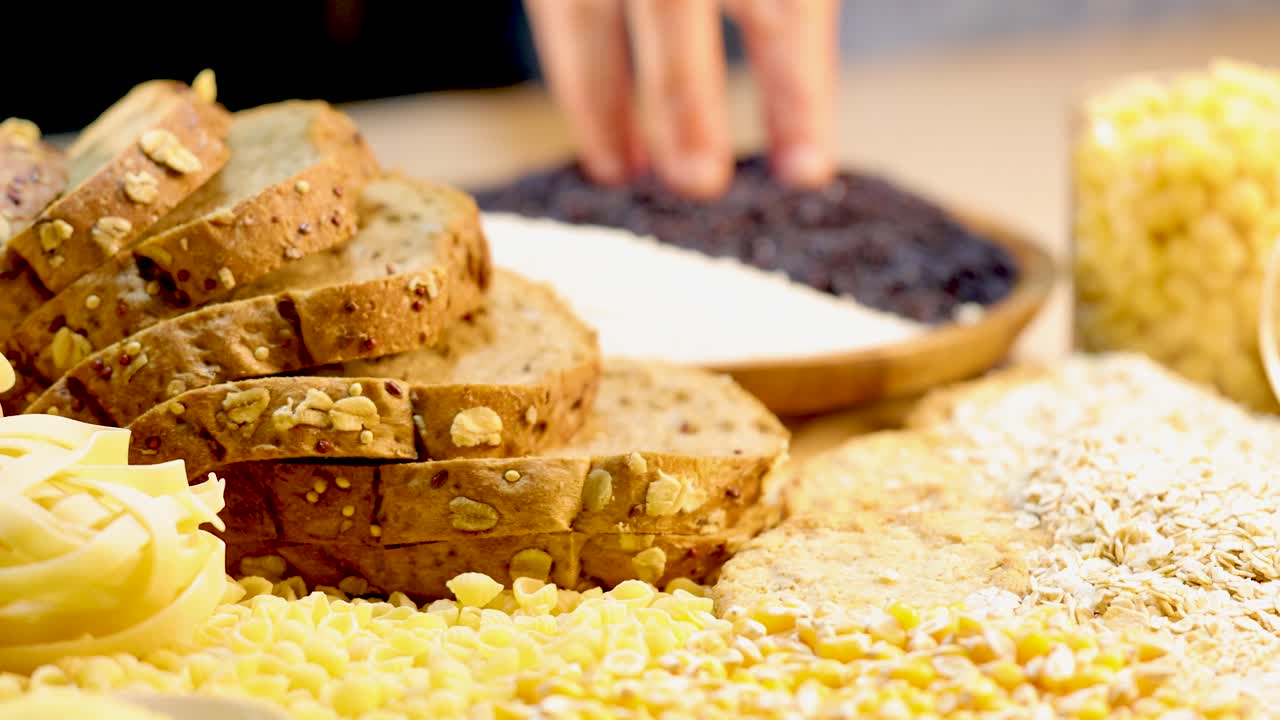 A close-up view of whole wheat bread, pasta, and grains with hands arranging ingredients in a bright kitchen setting