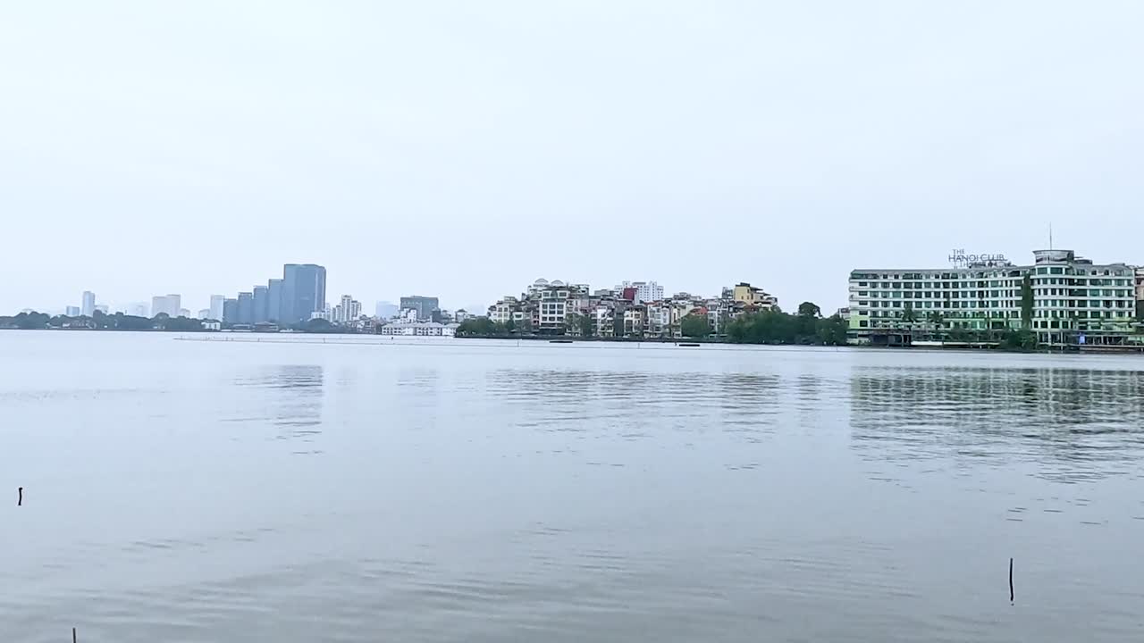 A tranquil view of urban buildings lining a calm waterfront under an overcast sky.