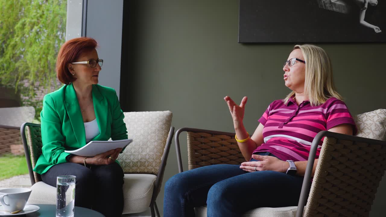 Two women engaged in conversation with a therapist, sitting on chairs in a bright, sunlit room, captured in slow motion