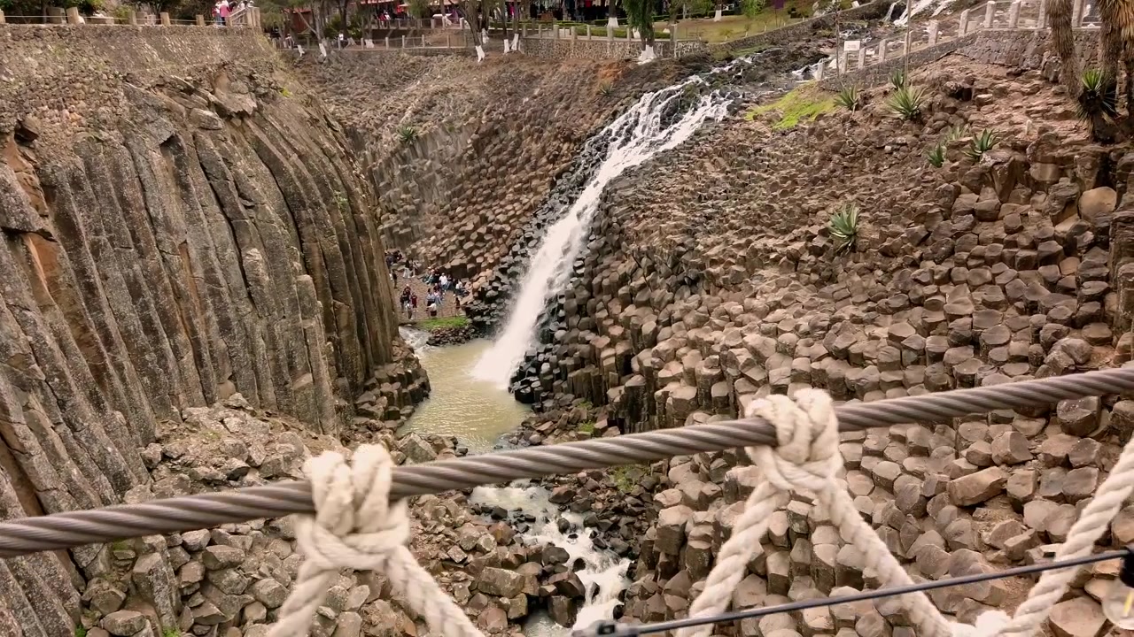 View from rope bridge overlooking waterfall flowing through dramatic basalt rock canyon surrounded by volcanic stone formations in Mexico