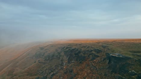 Vista-Aérea-Por-Drones-De-Las-Nieblas-Matutinas-Moviéndose-Lentamente-Sobre-Las-Escarpadas-Cimas-De-Las-Colinas-Peninas,-En-Una-Mañana-Nublada,-Colinas-Doradas-Y-Hermosos-Acantilados-Rocosos-Y-Páramos