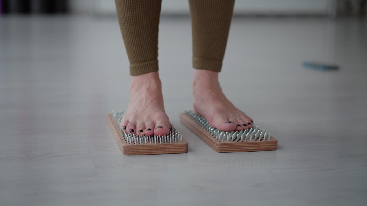 Leg view yoga specialist on brown leggings standing on wooden nail boards focusing on balance mindfulness pressure point therapy indoors in minimalist wellness studio under soft ambient light