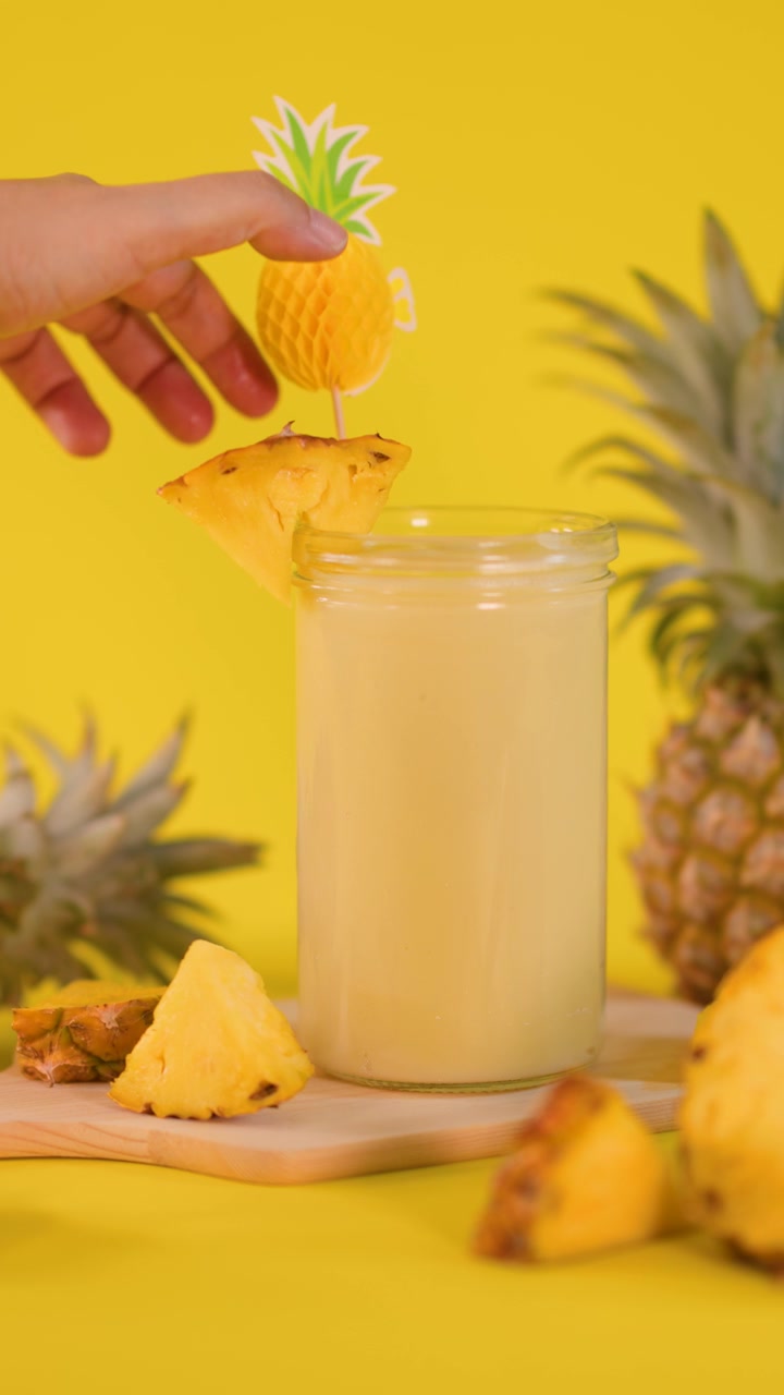 Una mano posiziona una fetta d'ananas e una decorazione di carta su un frullato in un ambiente da studio luminoso, tropicale e giallo