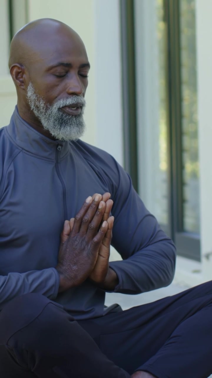 Vertical video: Lowering hands from brow, man in gray top and dark pants praying beside glass door