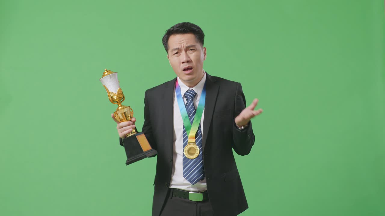 금메달을  슈트와 타이를 입은 아시안 사업가가 스튜디오의 녹색 스크린 배경에서 트로피를 의심스럽게 바라보고 있습니다.