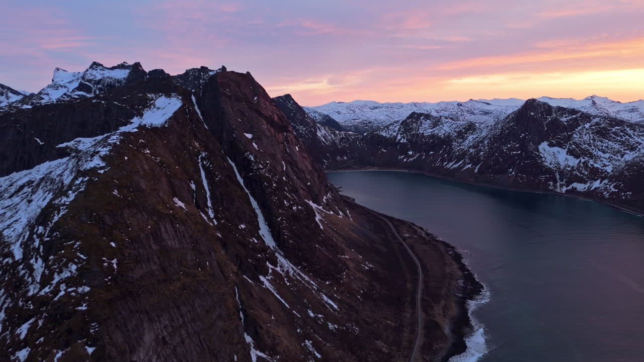 Maestosa catena montuosa al tramonto con cime innevate e una strada costiera tortuosa.