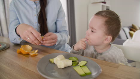 unrecognizable mom feeding her little son with clementine while sitting together at table in living room 1