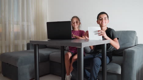 young boy finishes writing assignment in living room and shows work to someone while his sister watches attentively beside him at table with laptop during homework session