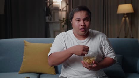 asian fat man watches tv and eats potato chips while sitting on a couch at home in the evening