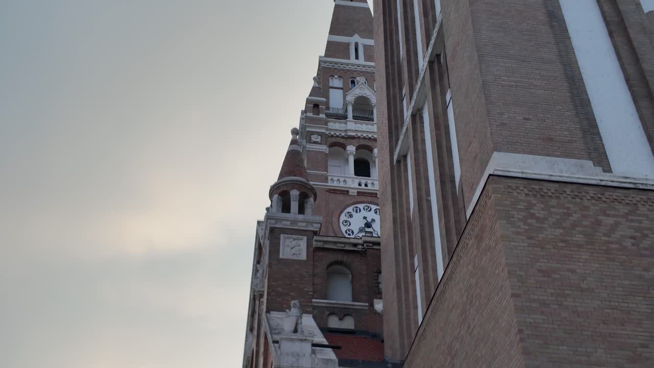 Upward view of the Votive Church in Szeged, focusing on the clock tower illuminated by warm sunset light